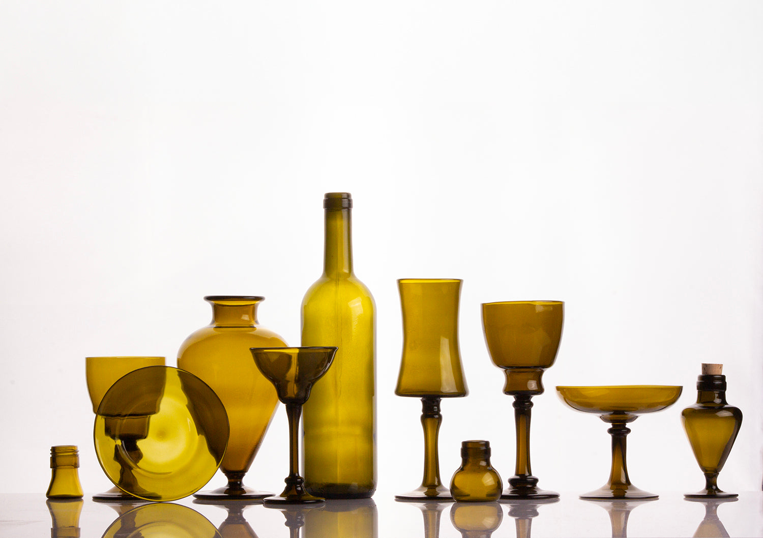 A collection of amber-yellow translucent glass vessels and drinkingware, including a tall bottle, goblets, a vase, and small containers, arranged on a reflective white surface against a white background. made from recycled glass.